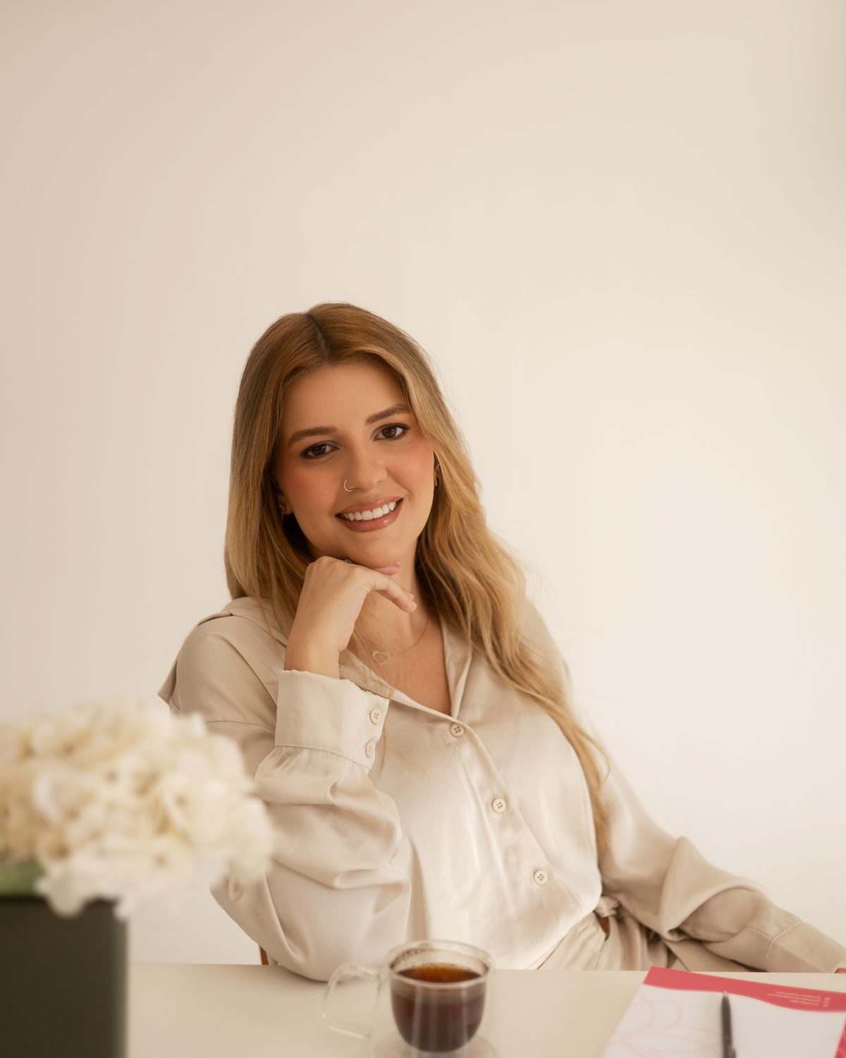 Lara Pansiere à mesa de trabalho, com caderno, xícara de café e um arranjo de flores brancas